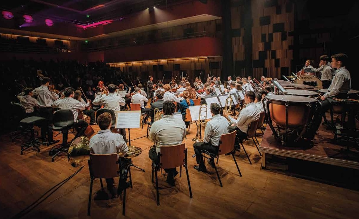 Orquestra jovem em apresentação no teatro, com plateia acompanhando o concerto